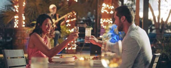 A smiling woman and man clink glasses at an outdoor restaurant table decorated with candles and string lights, while a musician plays guitar in the background.