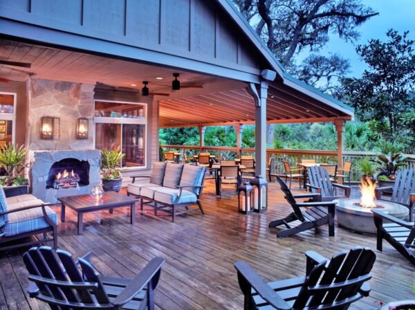 A spacious, covered wooden patio with cushioned seating, a stone fireplace, a fire pit, Adirondack chairs, potted plants, and lanterns, overlooking greenery at dusk.