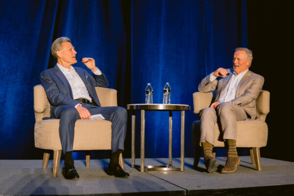 Two men in suits sit on armchairs on a stage with a blue curtain backdrop, speaking into microphones. A small round table with two water bottles is between them. Both men appear engaged in conversation.