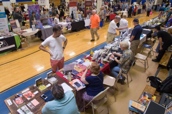 People converse and browse at a busy indoor book fair. Tables are covered with books, flyers, and displays. Authors and vendors sit behind the tables, while attendees walk, chat, and explore the event.