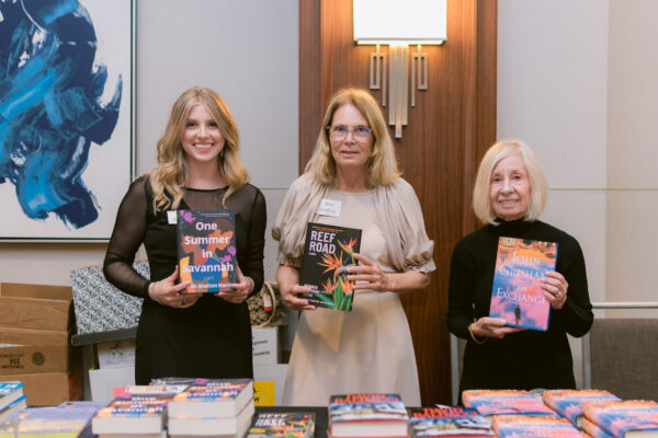 Three women stand behind a table of books, each holding up a novel and smiling at the camera. The setting appears to be indoors with a modern background, including a painting and a sconce.