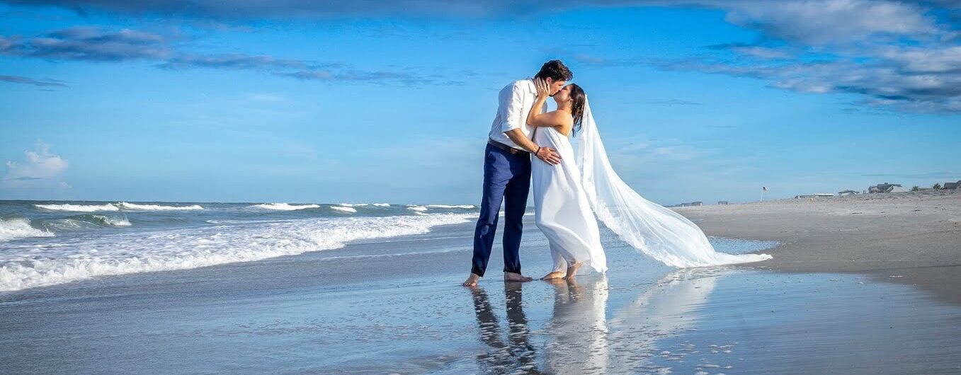 A bride and groom kiss on a sandy beach, reflected in wet sand. The brides veil flows in the wind, and the sky is bright blue with scattered clouds. Waves roll in beside them.