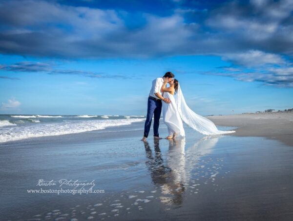 A bride and groom kiss on a sandy beach, reflected in wet sand. The brides veil flows in the wind, and the sky is bright blue with scattered clouds. Waves roll in beside them.