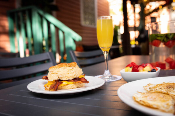 A breakfast scene on an outdoor table with a biscuit sandwich filled with bacon and cheese, a glass of orange drink, a bowl of mixed fruit, and a plate with fried eggs. Sunlight filters through trees in the background.