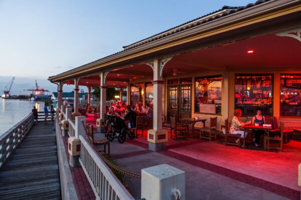 A waterfront restaurant at sunset with people dining inside and outside on a covered patio, illuminated by warm red lighting, next to a wooden dock and calm water.