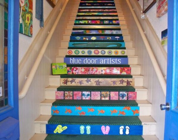 A staircase with colorful, hand-painted risers featuring fish, hearts, faces, and patterns, leading up to a landing with a sign that reads blue door artists.” A small “open” sign sits on one of the steps.