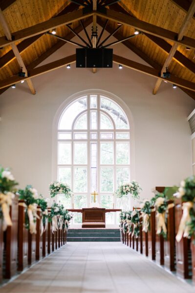 A bright, empty church interior with wooden pews decorated with white flowers and ribbons, leading to an altar adorned with floral arrangements and a gold cross, beneath a large arched window.