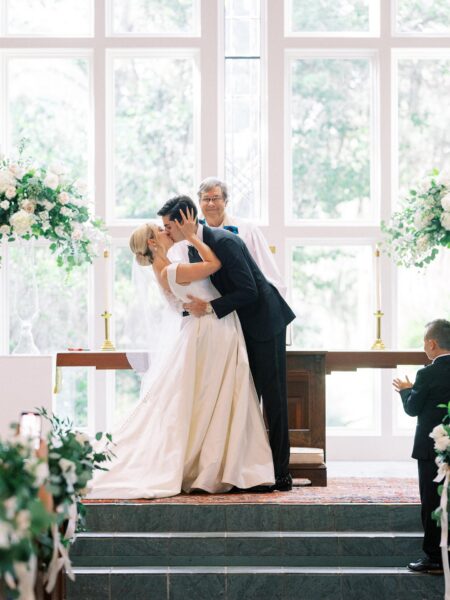 A bride and groom kiss at the altar during their wedding ceremony, with a smiling officiant behind them. A young boy stands nearby, clapping. The scene is set in a bright chapel decorated with flowers.