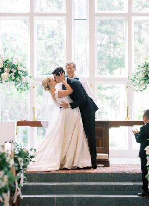 A bride and groom share a kiss at the altar during their wedding ceremony, with a smiling officiant behind them and a ring bearer clapping nearby. The setting is bright with large windows and floral arrangements.