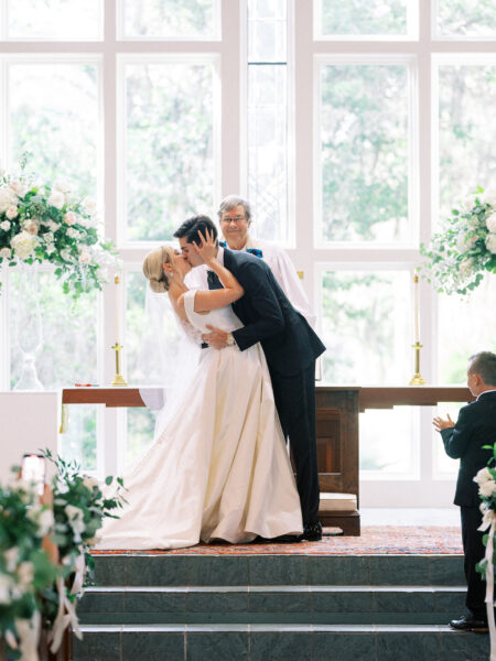 A bride and groom share a kiss at the altar during their wedding ceremony, as an officiant smiles behind them. The scene is set in a bright chapel with large windows and white floral arrangements. A young boy stands nearby, clapping.