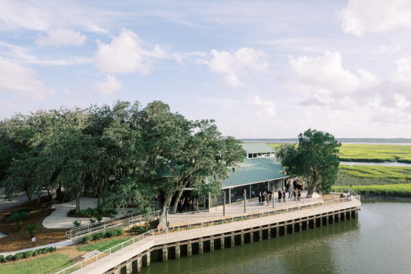 A large, open pavilion with a green roof sits on the edge of a river, surrounded by trees and marshland under a partly cloudy sky. Groups of people are gathered under the pavilion.