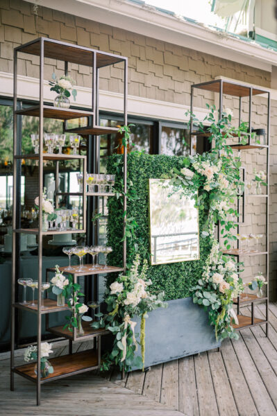 A decorative bar setup with metal and wood shelves holding glassware, lush greenery, and white flowers. A large framed mirror sign is centered on a green hedge backdrop. The scene is outdoors on a wooden deck.