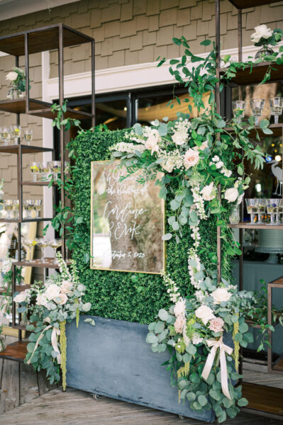 A decorative wedding welcome sign with calligraphy stands amid lush greenery and white florals, framed by leafy arrangements and surrounded by shelves holding glassware.