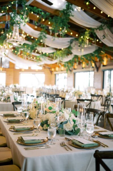Elegant wedding reception setup with white linens, floral centerpieces, glassware, and greenery hanging from the ceiling with string lights in a warmly lit, rustic venue.