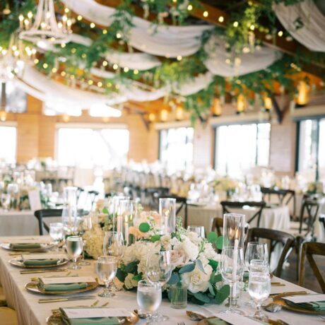 Elegant wedding reception setup with white linens, floral centerpieces, glassware, and greenery hanging from the ceiling with string lights in a warmly lit, rustic venue.