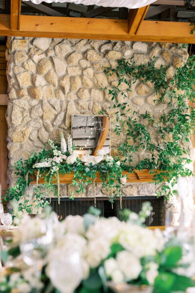 A stone fireplace decorated with lush green vines and white flowers, featuring a small sign with cursive writing on the wooden mantel. The foreground is softly blurred with more greenery and floral arrangements.