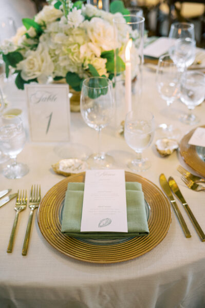 A formal table setting with gold-rimmed plates, gold cutlery, a green napkin, a printed menu, water and wine glasses, a white floral centerpiece, a lit candle, and a Table 1 card on a cream tablecloth.