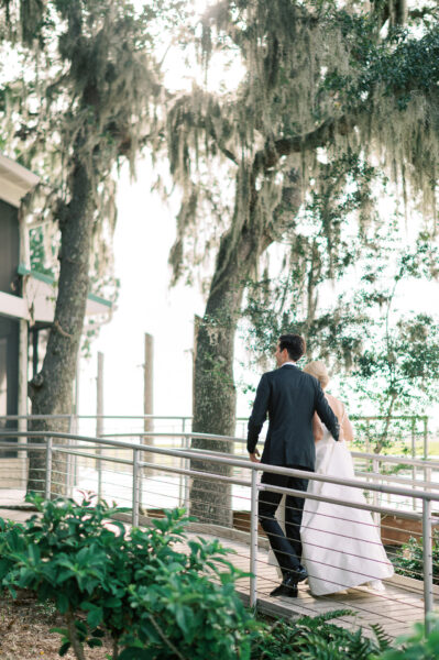 A couple, dressed in formal wedding attire, walks arm in arm along a wooden path surrounded by lush greenery and trees draped with Spanish moss, with sunlight filtering through the branches.