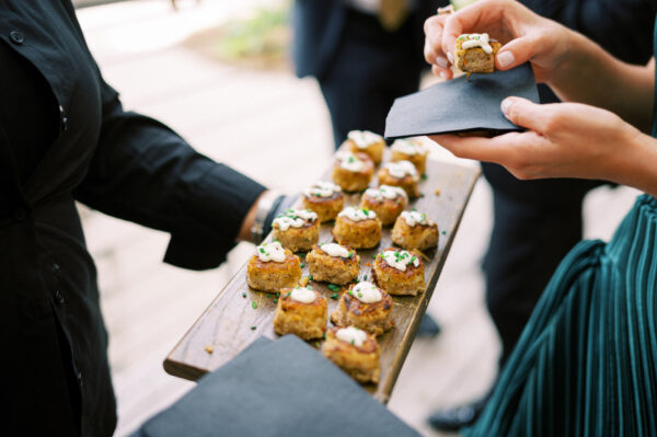 A person serves a wooden tray of bite-sized appetizers with a creamy topping to guests at an event. Another person takes one piece using a black napkin. The scene is lively and elegant.