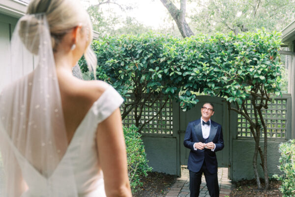 A bride in a white dress and veil stands facing a smiling man in a tuxedo, who stands under a leafy archway in a garden setting.