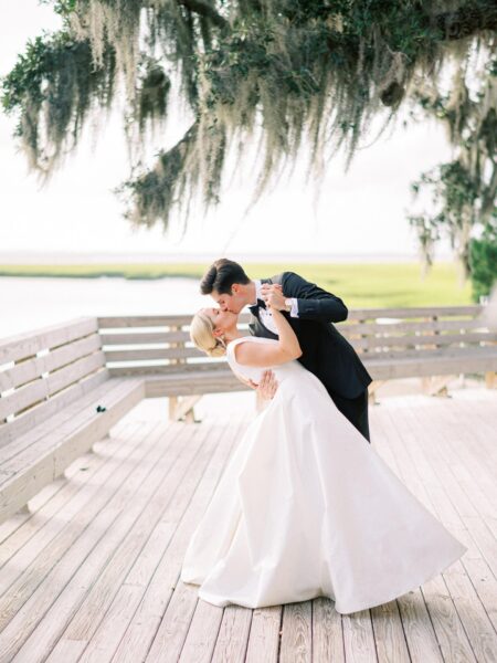A bride and groom share a romantic dip and kiss on a wooden deck under a tree, overlooking a scenic outdoor landscape.