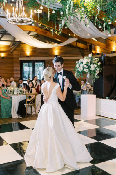 A bride and groom share their first dance on a black-and-white checkered floor in a decorated reception hall, with guests watching and greenery hanging from the ceiling.