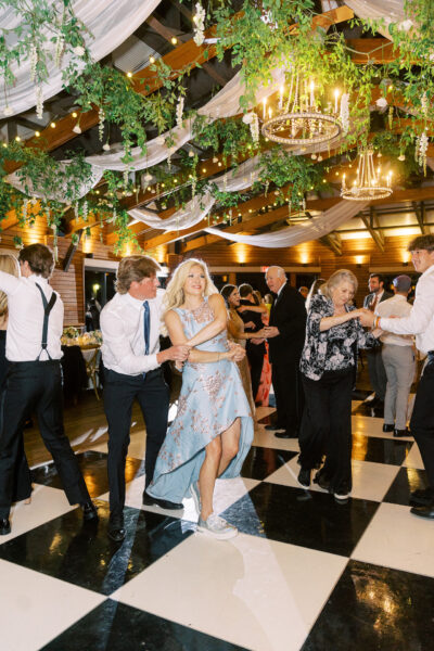 People dressed formally dance energetically on a black-and-white checkered floor under hanging greenery and chandeliers at a festive indoor event, with one woman in a blue dress smiling in the center.