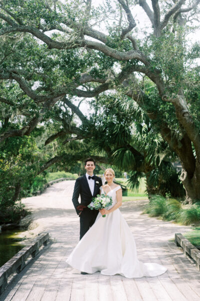 A bride and groom stand together on a wooden bridge under large, leafy trees. The bride wears a white dress and holds a bouquet; the groom wears a black tuxedo. Sunlight filters through the greenery behind them.