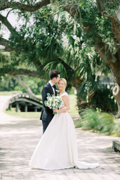 A bride in a white gown holds a bouquet and smiles while the groom, in a black suit, kisses her on the forehead. They stand outdoors on a sunlit path beneath lush green trees.