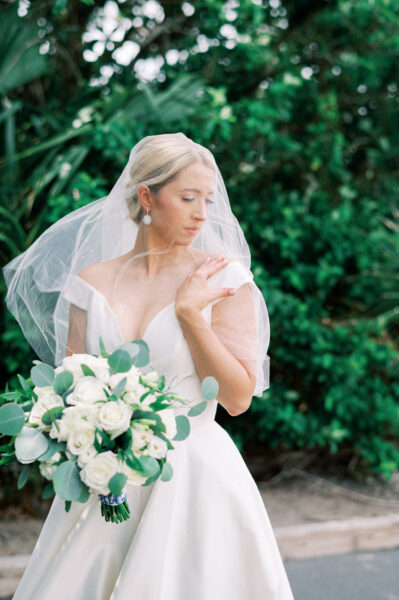 A bride in a white dress and veil holds a bouquet of white roses and greenery, standing outdoors with lush green foliage in the background. She looks down and gently touches her shoulder.