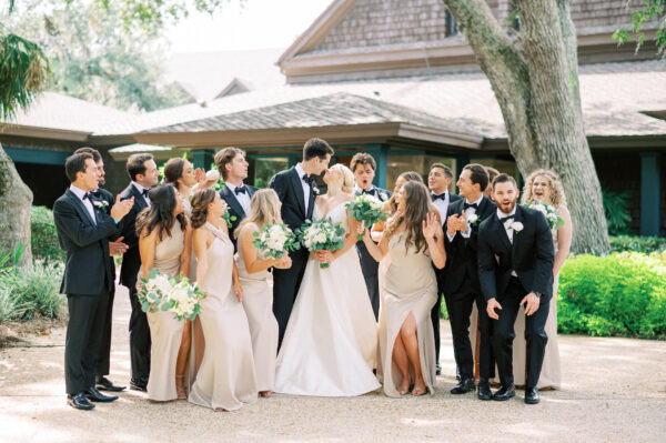 A wedding party of twelve people in formal attire, including the bride in white and bridesmaids in light dresses, stands outside smiling and laughing together in front of a house surrounded by greenery.