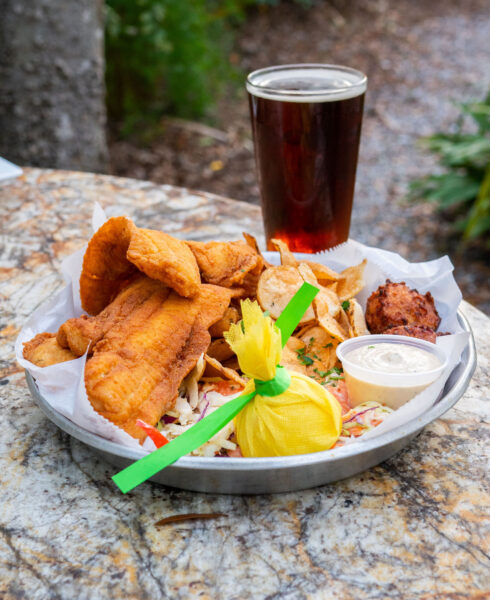 A plate of fried fish, potato chips, hush puppy, coleslaw, a lemon wrapped in yellow cloth, and a cup of dipping sauce sits on a stone table. A pint of dark beer is placed behind the plate. Outdoor setting with greenery in background.