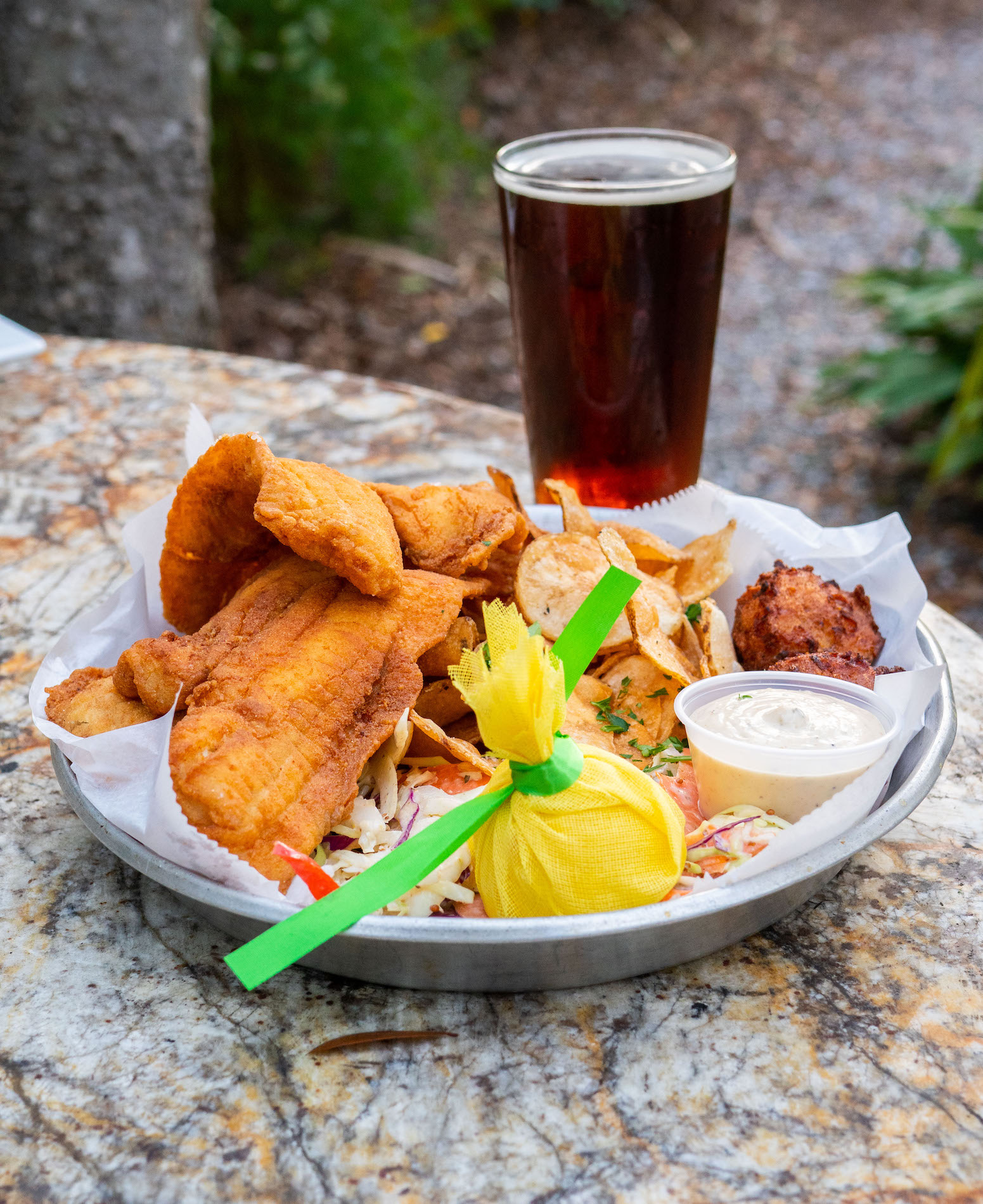 A plate of fried fish, potato chips, hush puppy, coleslaw, a lemon wrapped in yellow cloth, and a cup of dipping sauce sits on a stone table. A pint of dark beer is placed behind the plate. Outdoor setting with greenery in background.