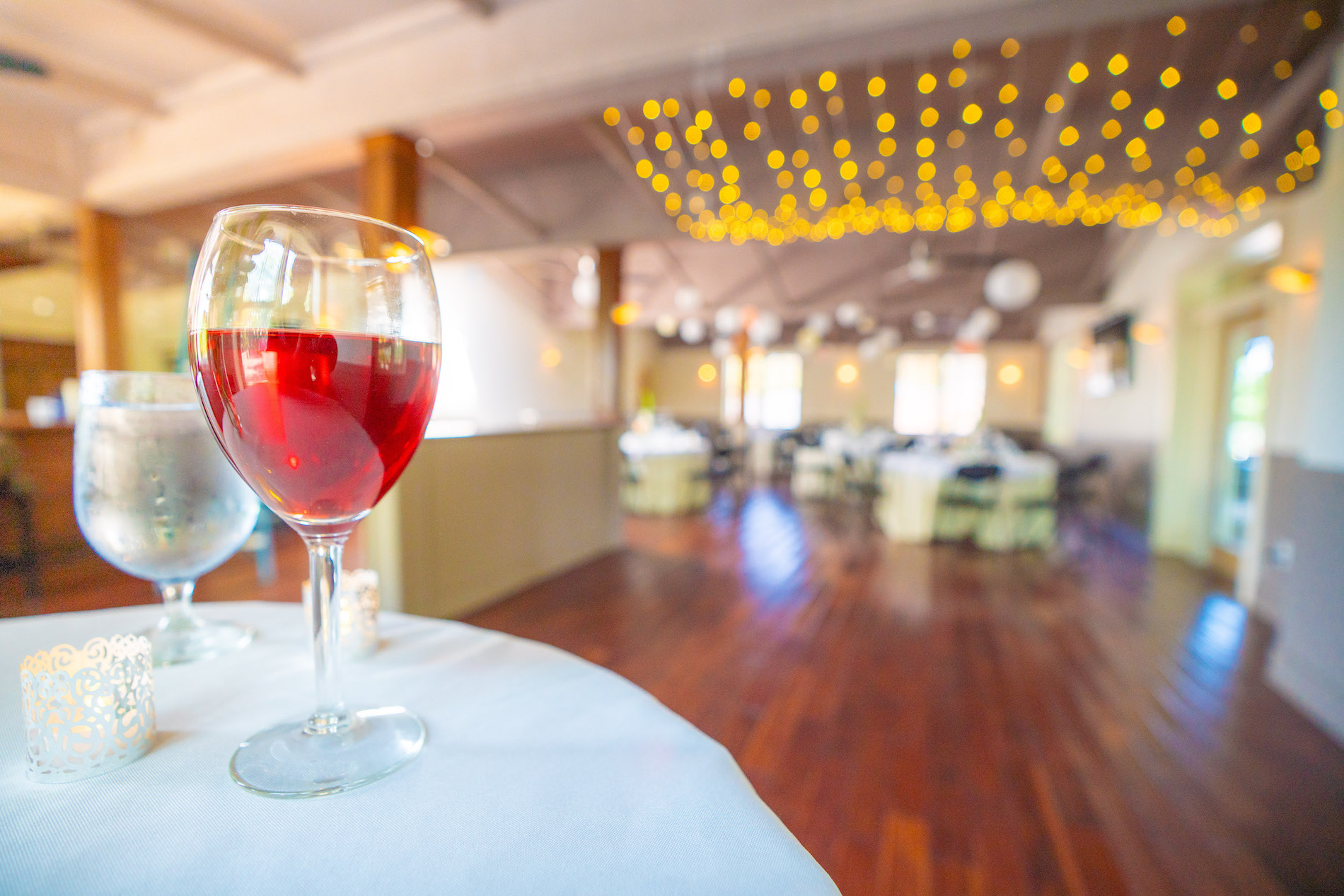 A glass of red wine and a glass of water sit on a table in a warmly lit banquet hall with wooden floors and hanging string lights, with round, set tables visible in the blurred background.