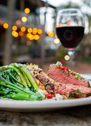 A plate of sliced medium-rare steak with sautéed broccolini and mashed potatoes sits on an outdoor table, with a glass of red wine and blurred string lights in the background.