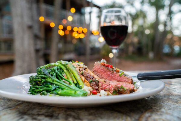 A plate of sliced medium-rare steak with sautéed broccolini and mashed potatoes sits on an outdoor table, with a glass of red wine and blurred string lights in the background.