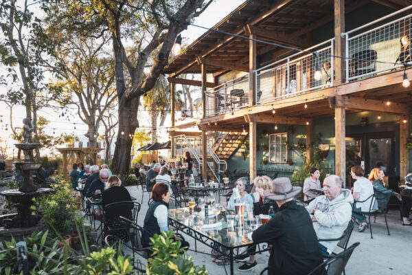 People sit and dine at outdoor tables on a patio surrounded by greenery and string lights, next to a two-story building with a balcony, during daylight. The atmosphere is lively and relaxed.