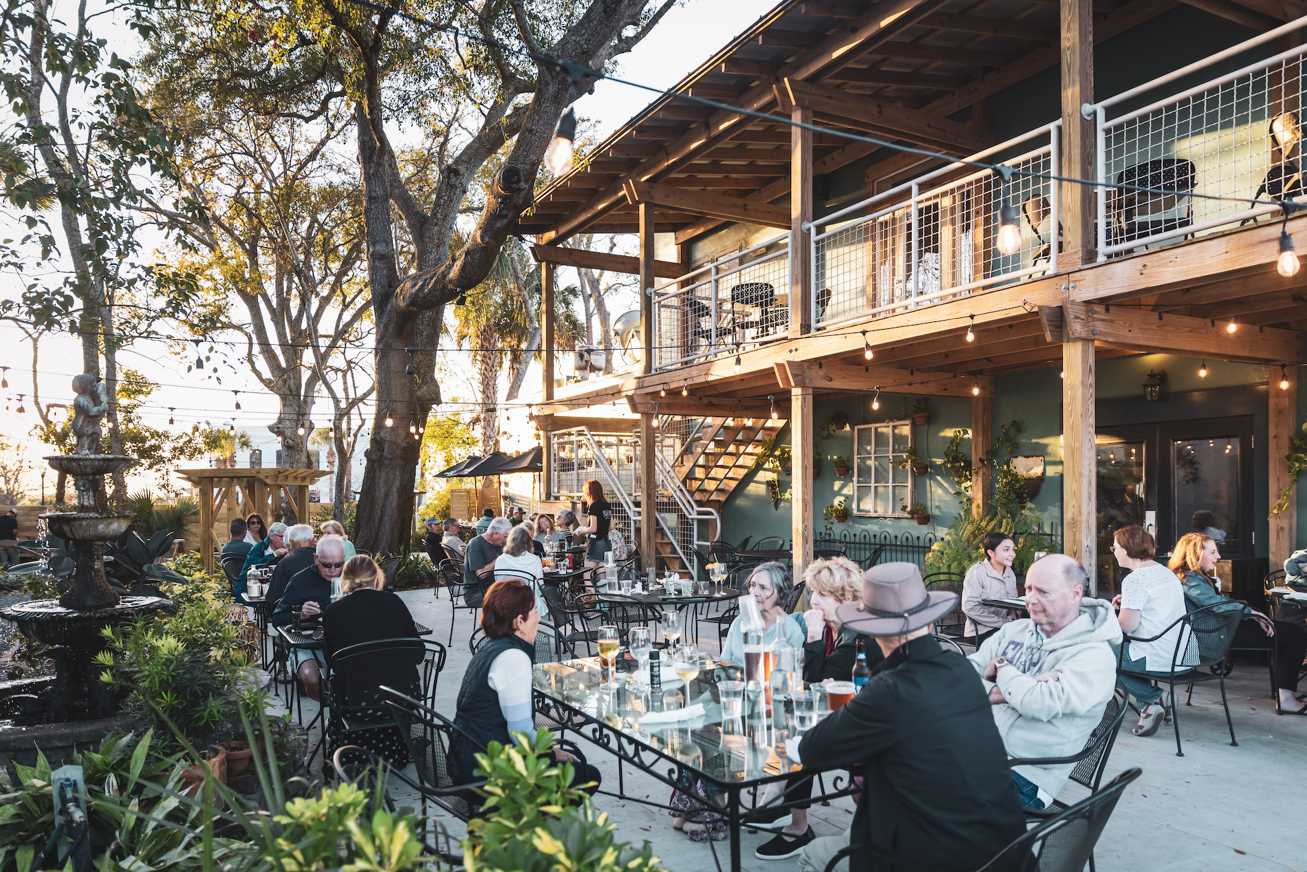 People sit and dine at outdoor tables on a patio surrounded by greenery and string lights, next to a two-story building with a balcony, during daylight. The atmosphere is lively and relaxed.