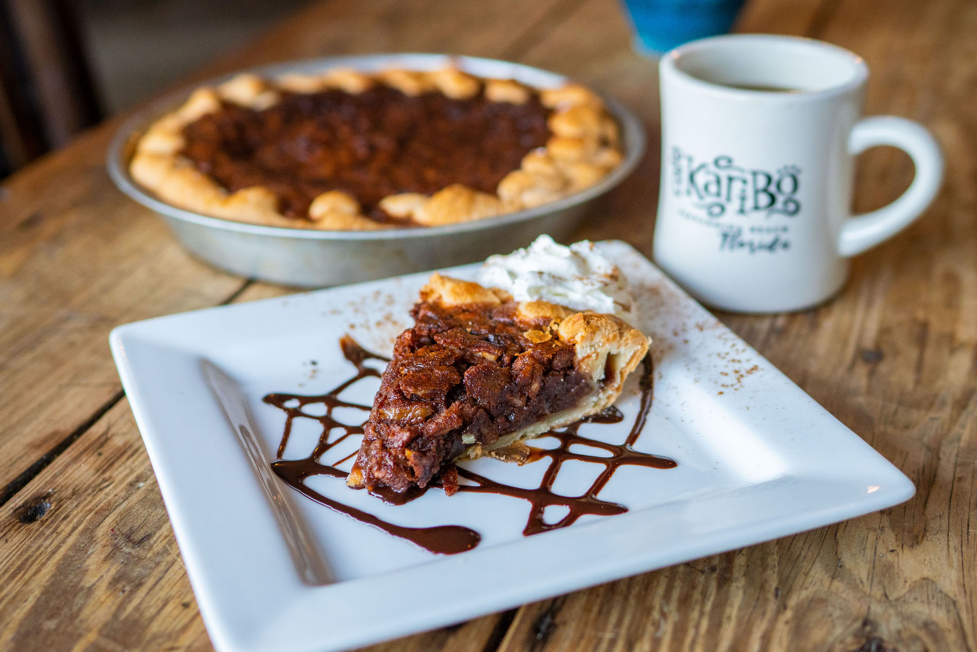 A slice of chocolate pie with whipped cream and chocolate drizzle sits on a white plate, with the rest of the pie and a mug of coffee in the background on a wooden table.