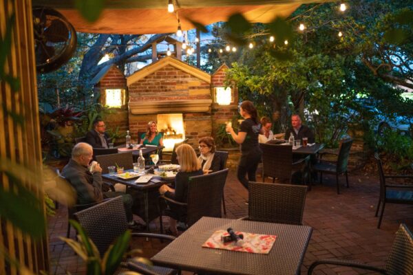 People dining at outdoor restaurant tables on a brick patio under string lights in the evening, with a fireplace in the background and a server walking between the tables.