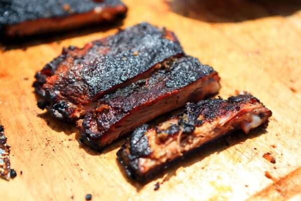 Three pieces of barbecued meat with a dark, charred crust are arranged on a wooden cutting board. The meat looks juicy and tender, with a smoky, caramelized exterior.