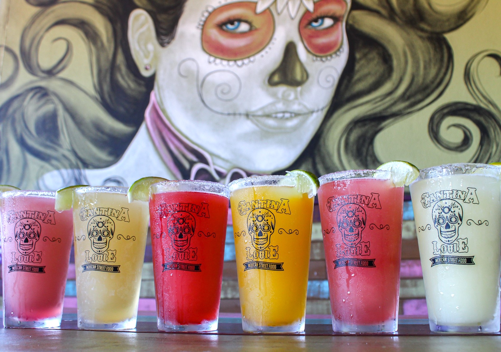 Seven colorful drinks with salted rims and lime wedges are lined up on a bar, with a mural of a womans face painted in a Day of the Dead style in the background.