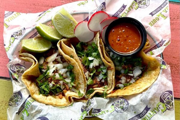 Three street-style tacos filled with seasoned meat, chopped onions, and cilantro, served with lime wedges, sliced radishes, and a small cup of red salsa on branded paper.