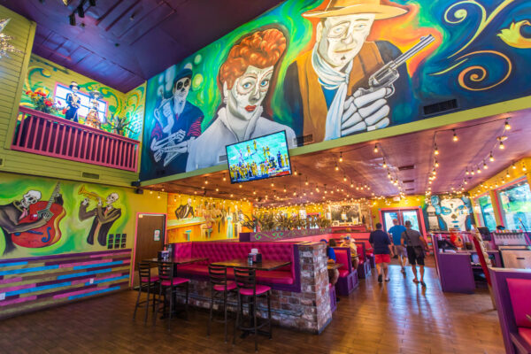 A vibrant restaurant interior with colorful murals of iconic figures on the walls, string lights hanging from the ceiling, pink booths, a TV, and a few people walking inside.