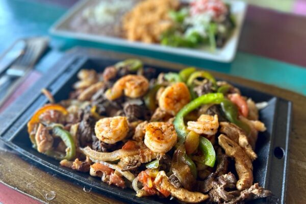 A sizzling platter of fajitas with shrimp, chicken, beef, bell peppers, and onions sits in focus, while a blurred plate of beans, rice, and salad is in the background.