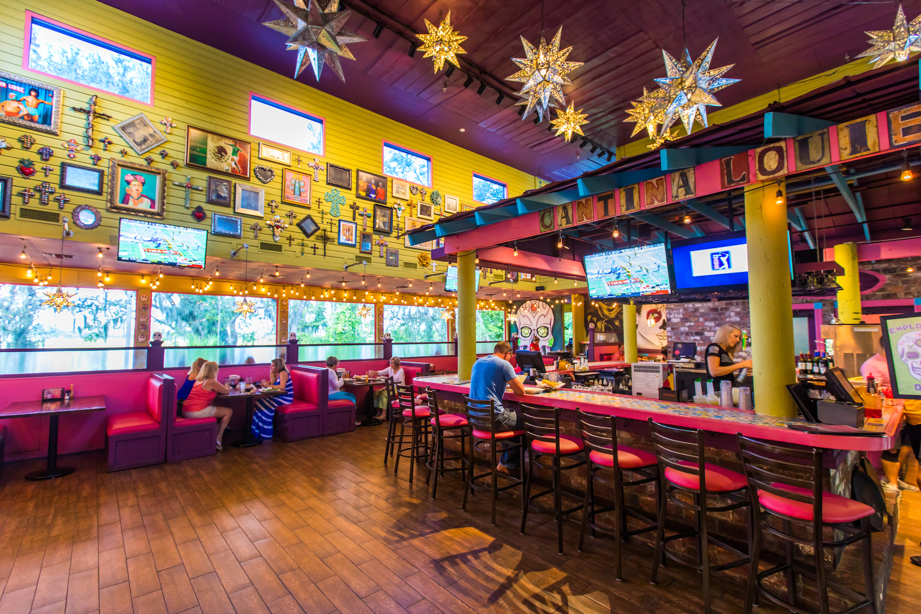 Colorful Mexican restaurant interior with star-shaped hanging lights, vibrant wall art, pink booths, and a bar with high chairs. A few patrons are seated, and large windows let in natural light.
