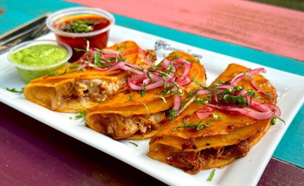 Three birria tacos filled with shredded meat and melted cheese, topped with pickled onions and herbs, served on a white plate with green and red dipping sauces on the side, on a colorful table.