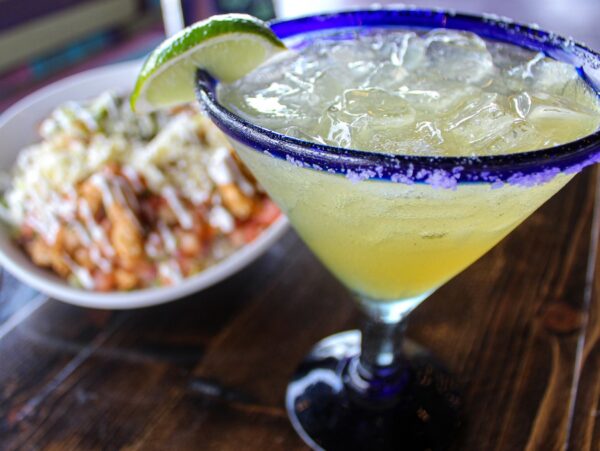 A close-up of a salted margarita glass with ice and a lime wedge, sitting on a wooden table. In the background, there is a blurred bowl of food topped with sauce and cheese.
