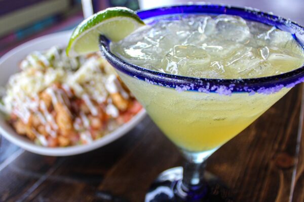 A close-up of a margarita with a salted rim and lime wedge, served in a glass on a wooden table, with a blurred dish of nachos topped with cheese and sauce in the background.