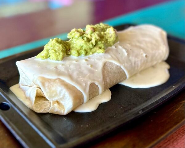 A chimichanga topped with a creamy white sauce and a scoop of guacamole, served on a black rectangular plate.